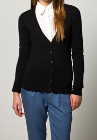 Svart kofta med V-ringning, långa ärmar och knäppning fram, bärs över en vit skjorta med krage. Strukturstickad material.