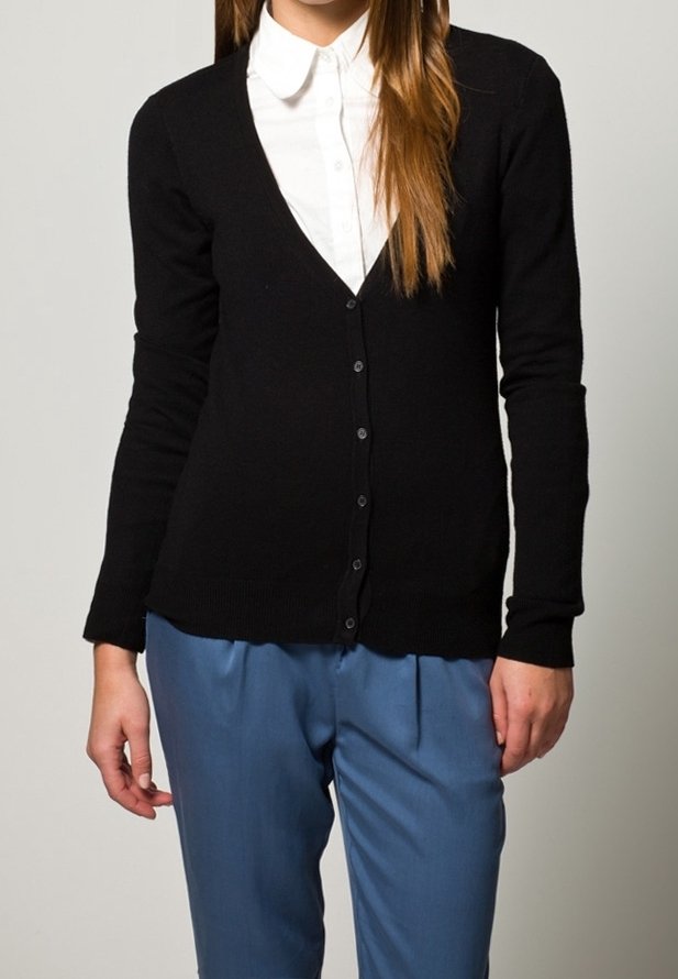 Svart kofta med V-ringning, långa ärmar och knäppning fram, bärs över en vit skjorta med krage. Strukturstickad material.