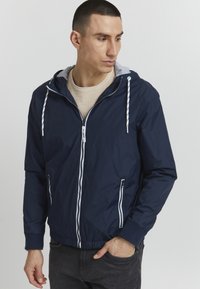 Chaqueta ligera de color azul marino con cremallera frontal, forro de capucha gris, cordones blancos y bolsillos laterales. Textura suave, puños elásticos.