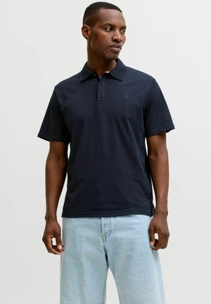 Férfi fekete rövid ujjú galléros pólóban és világoskék farmerben, kezét lazán tartva, oldalra nézve.