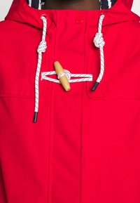 Chaqueta de lluvia roja con capucha, con tejido texturizado, cordones blancos anudados, un cierre de madera y un bolsillo en el lado izquierdo.