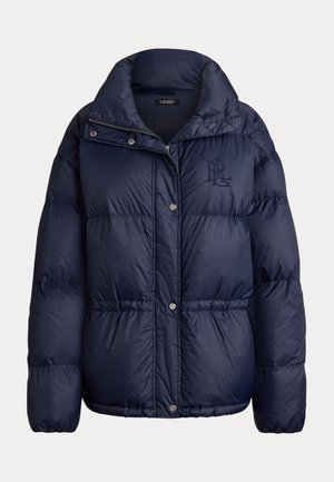 Femme portant une veste matelassée bleu foncé, un jean bleu et des chaussures marron, tenant un sac à main marron, debout dans un environnement intérieur neutre.