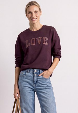 Kvinde smilende iført en mørkerød sweatshirt med teksten "LOVE", lyseblå jeans og holder en brun håndtaskerem i venstre hånd.