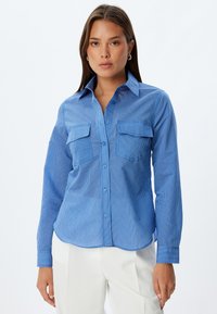Camisa de botones a rayas azules con manga larga, dos bolsillos en el pecho y cuello extendido; hecha de una tela lisa y ligera.