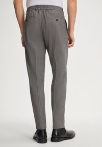 Uomo in pantaloni grigi su misura con tasche posteriori con bottone e scarpe eleganti in pelle nera, in piedi di fronte a uno sfondo neutro.