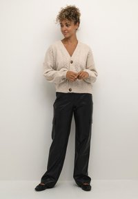 Beige strikket cardigan med V-hals, lange ærmer og knaplukning. Har et zigzag-mønster og blødt, tekstureret stof. Kombineret med løse, sorte læderbukser.