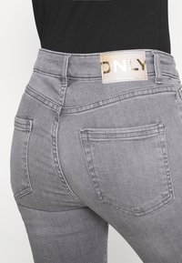 Jeans de denim cinza com um design ajustado. Apresentam um patch dourado com o logótipo na cintura e dois bolsos traseiros com detalhes em costura.