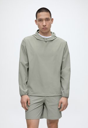 Mladý muž v svetlosivej mikine s kapucňou a zodpovedajúcich šortkách, stojaci s neutrálnym výrazom tváre na jednoduchom bielom pozadí.