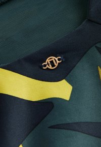 Grön och gul mönstrad tyg med en texturerad nätpanel, med en detalj i guldhardware formad som en loop och en rektangel.