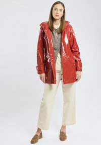 Rode, glanzende, waterdichte jas met capuchon, voorkeursrits en drukknoopsluitingen. Gecombineerd met een grijze trui, crèmekleurige broek en schoenen met dierenprint.