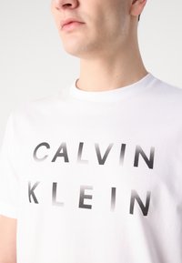 White cotton t-shirt featuring a gradient black text print "CALVIN KLEIN" across the chest. Rounded neck and short sleeves.