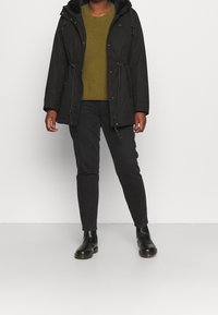 Parka noire avec capuche, fermeture éclair et détails à cordon, portée sur un pull vert olive et un jean noir ; des bottes noires complètent l'ensemble.