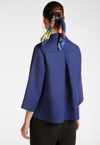 Blusa di lino blu con maniche a tre quarti, vestibilità morbida, dettaglio plissè sul retro e una sciarpa in seta colorata annodata sul retro.