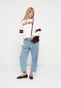 Biały skrócony sweatshirt z czerwonym logo "GAP NYC", niebieskie luźne dżinsy i błyszczące ciemno burgundowe baleriny; trzymając małą czarną torebkę na ramię.