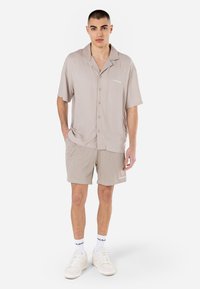 Chemise beige à manches courtes avec un col et des boutons, associée à un short en maille beige clair avec un accent logo. Portée avec des baskets blanches.