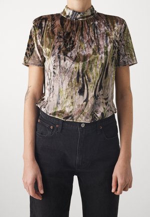Personne portant un chemisier à manches courtes, col montant, avec un motif abstrait marron, vert et noir, rentré dans un jean noir taille haute.