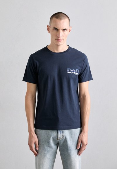 Navy blue cotton t-shirt featuring a graphic "DAD" print in light blue on the left side. Short sleeves and crew neckline, paired with light denim jeans.