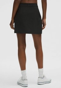Falda de algodón negra con corte recto, cinturilla y dos bolsillos traseros; combinada con zapatos y calcetines blancos. Textura suave y diseño minimalista.