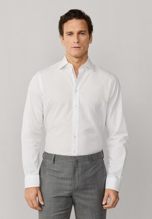 Mann trägt ein weißes, langärmeliges Hemd mit Knöpfen und graue, maßgeschneiderte Hose, steht vor einem schlichten hellgrauen Hintergrund.