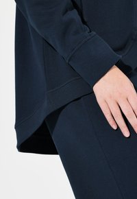 Marinblå sweatshirt i mjuk bomullsblandning, med avslappnad passform, ribbade ärmslut och rundad nederkant med sidokilar. Närbild av hand som vilar på tyget.