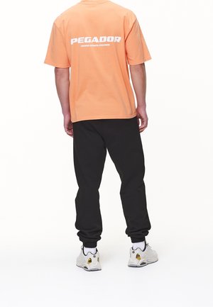 Personne portant un t-shirt pêche avec le texte « PEGADOR » dans le dos, un pantalon de jogging noir, des chaussettes blanches et des baskets blanches, debout de dos.