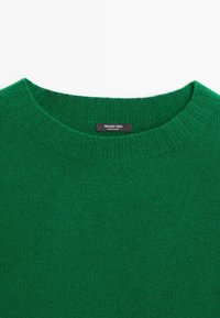 Pull en maille vert à col rond côtelé avec une étiquette noire portant la mention « Massimo Dutti Made in China ».