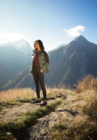 Grønn pufferjakke med svarte detaljer, maroon topp og svarte leggings. Står på en sti med fjell og gress i bakgrunnen.
