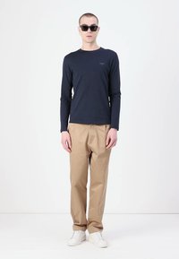 T-shirt a maniche lunghe blu abbinato a pantaloni beige e sneakers bianche. Occhiali da sole indossati; design minimalista con una vestibilità casual.