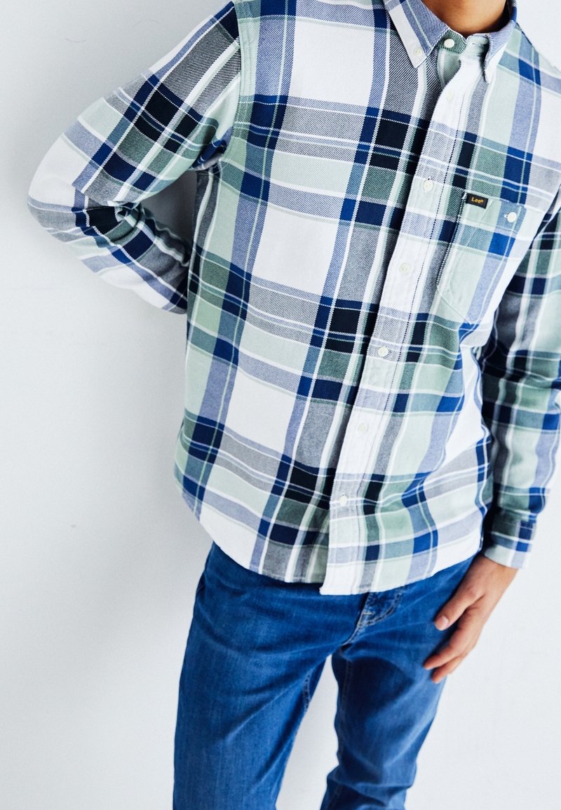 Personne portant une chemise à carreaux bleue, blanche et verte à manches longues et un jean bleu, debout avec une main sur la hanche.