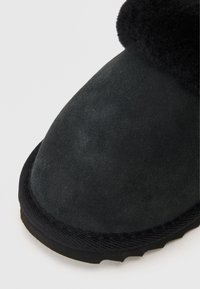 Μαύρο σουέτ παντόφλα με στρογγυλεμένη μύτη, διαθέτοντας ένα μαλακό, υφασμάτινο εσωτερικό και μια ανθεκτική καουτσούκ σόλα με διακριτό μοτίβο πέλματος.