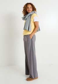 Chaleco de punto amarillo y esponjoso sobre una camiseta blanca, pantalones grises de pierna ancha con franjas laterales y una bufanda gris claro, combinados con zapatillas marrones con estampado animal.