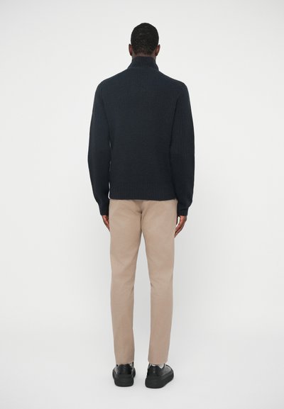 Musta ribattu neulepusero korkealla kauluksella, käytettynä beigellä slim-fit-housuilla ja mustilla lenkkareilla, nähtynä takaa.