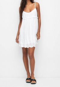 Mujer vistiendo un vestido blanco sin mangas hasta la rodilla con detalles de encaje y sandalias negras de tipo deslizante, de pie frente a un fondo blanco liso.