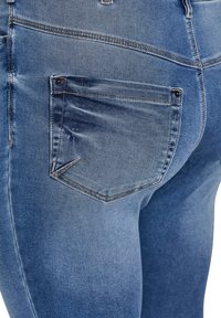 Plave traperice uskog kroja, s džepom na stražnjoj strani opremljenim crnim metalnim detaljima i blago izblijedjelim teksturom tkanine.