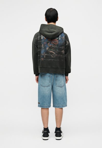 Σκούρο πράσινο φούτερ με κουκούλα και γραφικό σχέδιο, συνδυασμένο με ανοιχτό μπλε τζιν σορτς. Μαύρα αθλητικά παπούτσια ολοκληρώνουν την εμφάνιση. Πλάγια όψη, απλό φόντο.