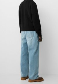 Person som bär ljusblå vida jeans, bruna skor och en svart stickad tröja, står mot en enfärgad vit bakgrund.