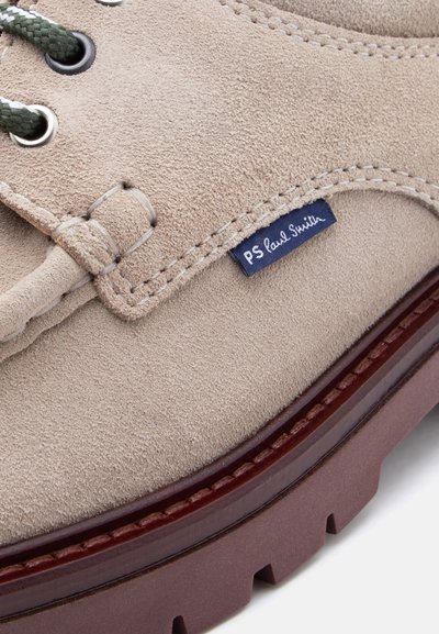 Gros plan sur une chaussure en daim beige avec des lacets verts et blancs, une couture détaillée, une semelle marron foncé, et une étiquette en tissu bleue "PS Paul Smith".