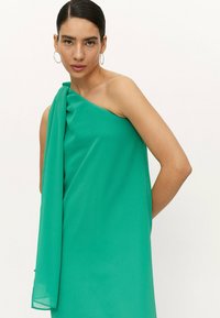 Vestido verde sin mangas confeccionado en un tejido ligero, con un acento drapeado en un lado y una textura suave y elegante.