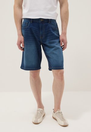 Mann trägt mittellange blaue Jeansshorts, weißes T-Shirt und weiße Sneaker und steht vor einfarbigem Hintergrund.