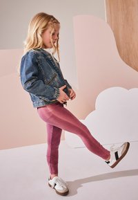 Jeans jakna z zadrgo, roza teksturirane legice in bele superge z rjavimi detajli. Ozadje prikazuje mehke pastelne oblike.