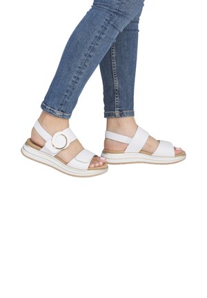 Feet wearing white open-toe sandals with thick straps and a gold buckle, paired with blue skinny jeans against a white background.