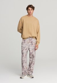 Felpa oversize beige con scollo rotondo abbinata a pantaloni grigio chiaro con motivo e sneaker grigie. I pantaloni presentano un design astratto e testurizzato.