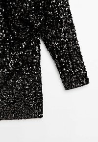 Vêtement noir à manches longues, densément couvert de sequins noirs brillants sur fond blanc.