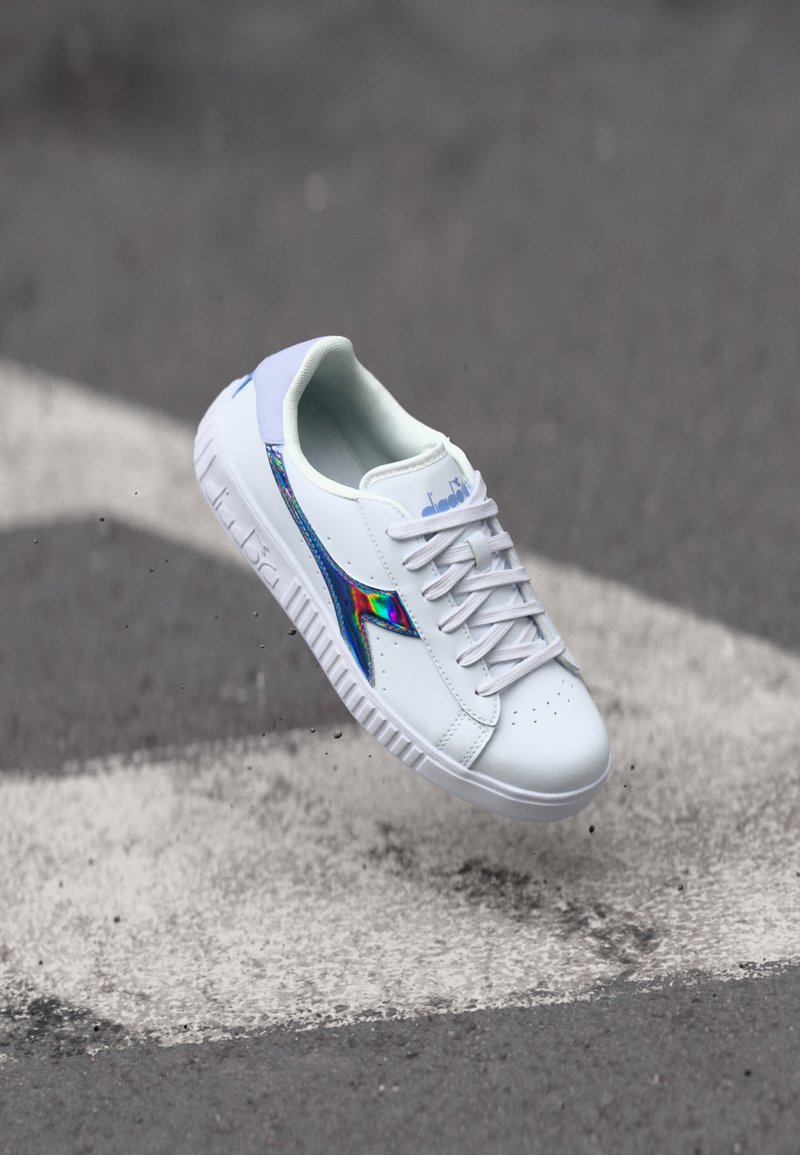 Fehér tornacipő sima szintetikus felsőrésszel, kék és irizáló oldalsó díszítéssel, fűzős kivitelben és mintás gumitalppal.