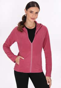 Różowy polarowy hoodie z przednim zamkiem, długimi rękawami i bocznymi kieszeniami. Miękka tekstura i luźny krój, zestawiony z czarną bluzką pod spodem.