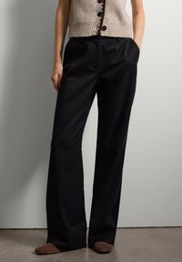 Pantalon noir sur mesure avec un design à jambe droite, présentant une texture lisse et des poches latérales, associé à un haut beige sans manches à côtes.