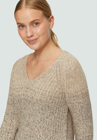 Beige gebreide trui met een V-hals, voorzien van een gestructureerd patroon en lange mouwen, die een zachte, casual uitstraling heeft.