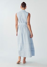 Šviesiai mėlynas, be rankovių maksi suknelė su diržu juosmenyje, sluoksniuotas dizainas ir apykaklė. Lygus audinys su švelniu tekstūru.