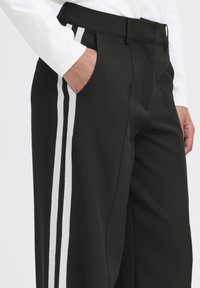 Pantaloni neri con strisce laterali bianche, caratterizzati da una vestibilità su misura, una texture liscia e tasche laterali. La mano dell'utente è appoggiata nella tasca.