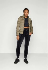 Femme portant une veste matelassée vert olive, un crop top noir, un pantalon de jogging noir, des chaussettes blanches et des baskets noires se tenant devant un fond uni.